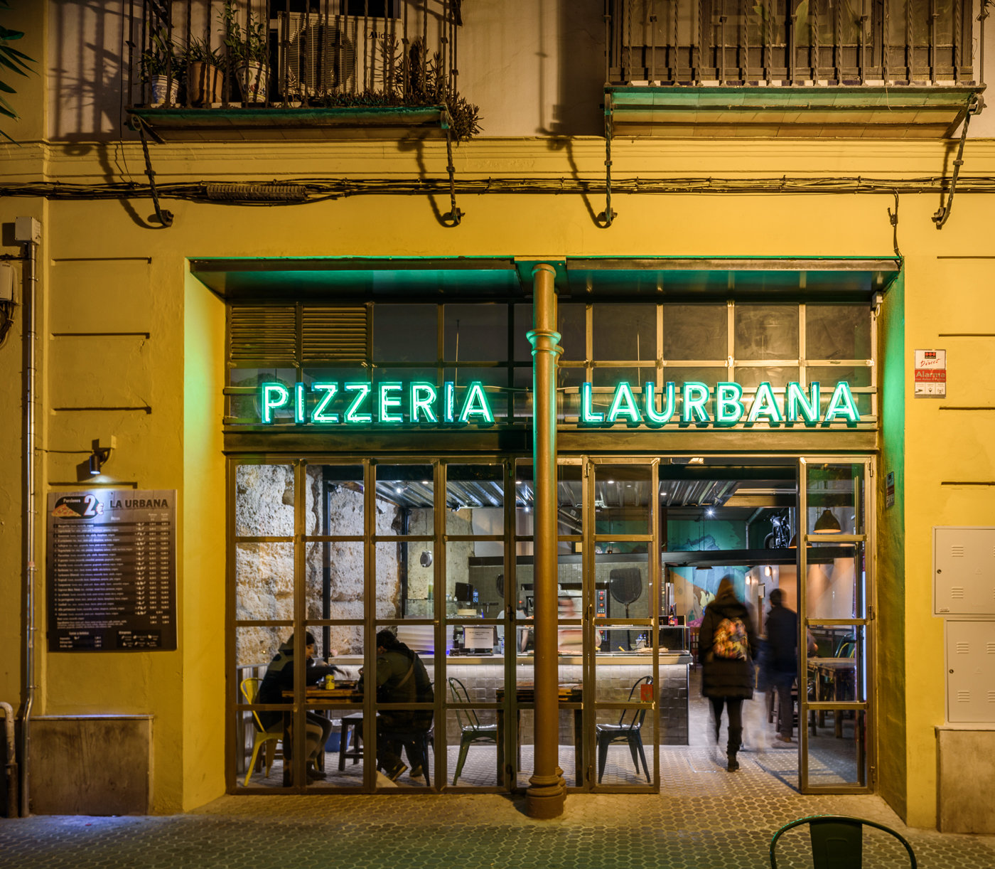 La pizzería de noche con el rótulo de neón encendido y clientes en las mesas