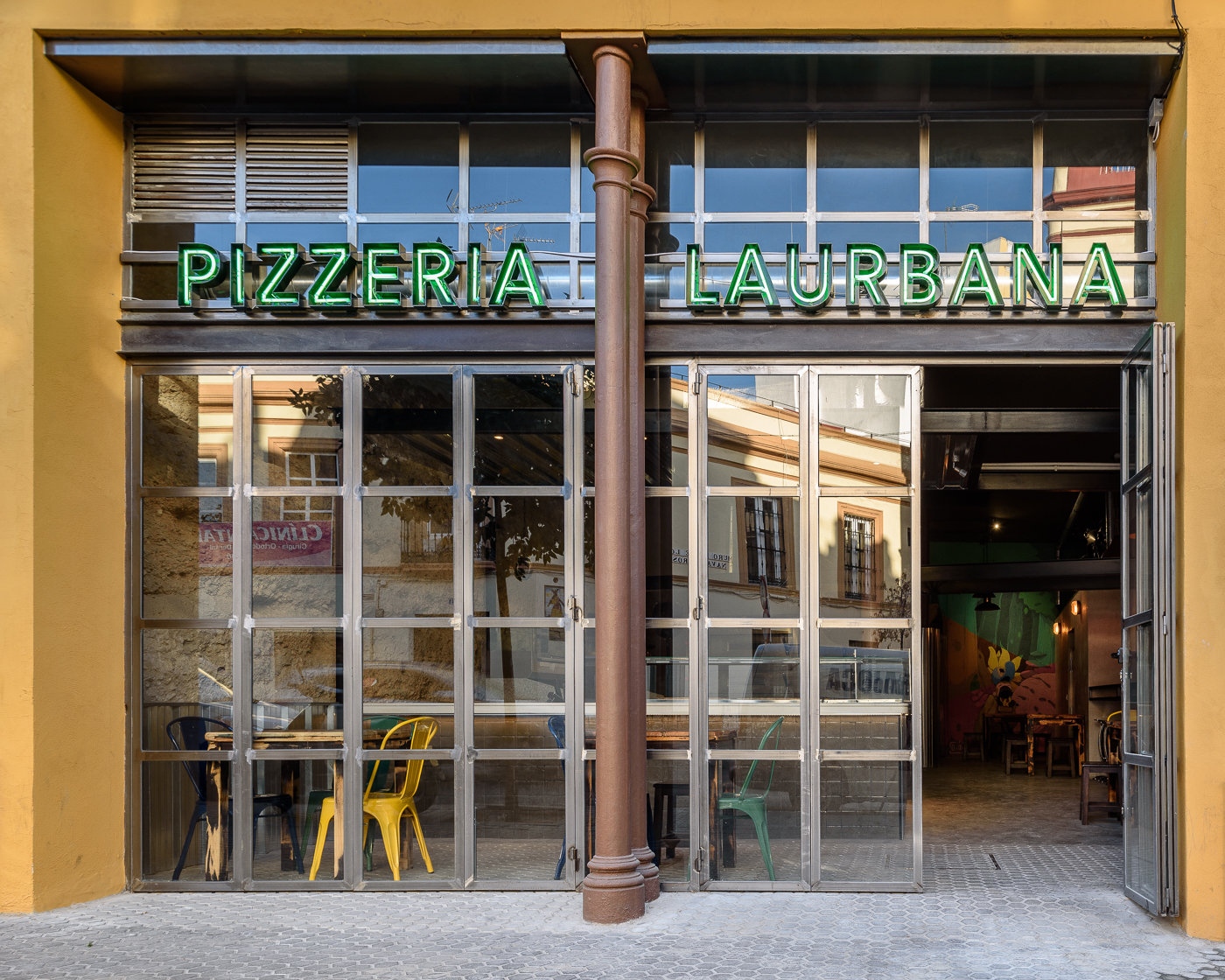 Fachada de la pizzería con una hoja abierta y las mesas del interior tras el vidrio