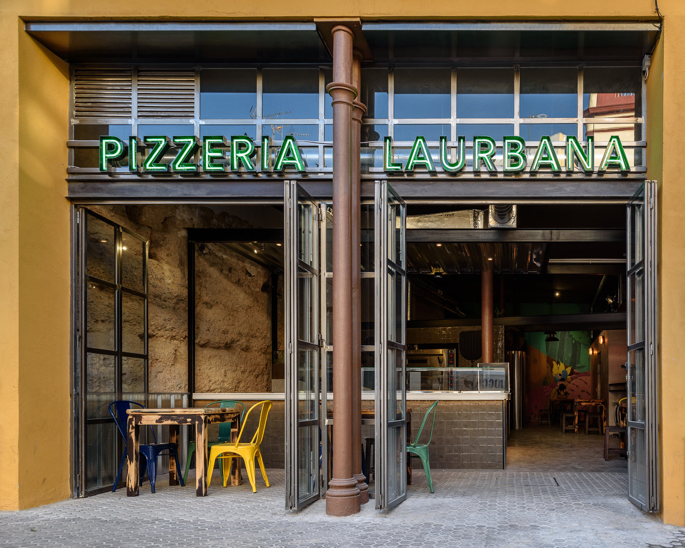 La pizzería con las puertas plegables abiertas y sillas de colores en la calle