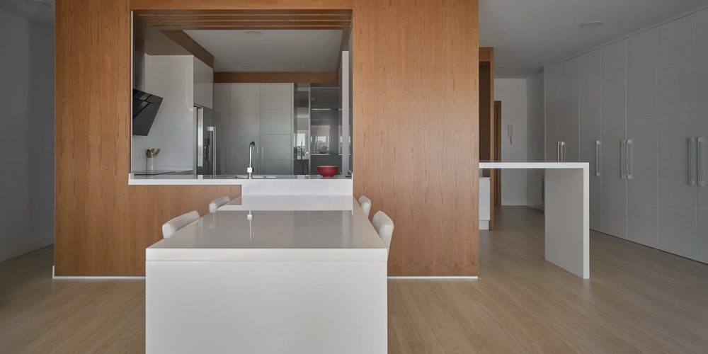 Cocina abierta por un hueco en el panelado de madera, con isla blanca delante