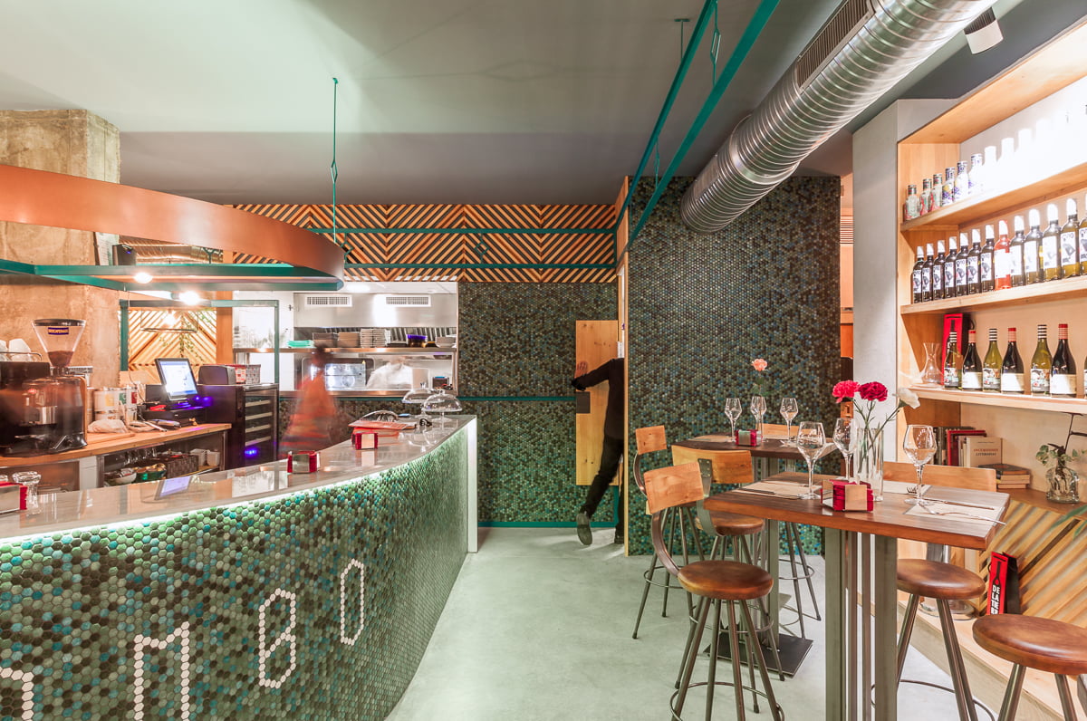 Barra curva de mosaico verde frente a la cocina vista, con mesas altas y vinos