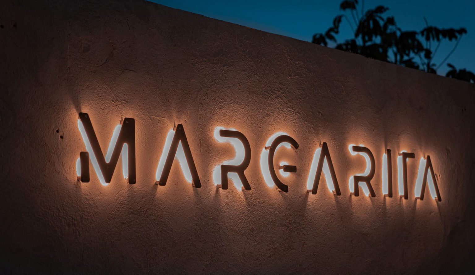 Letras retroiluminadas de Margarita sobre un muro enfoscado al anochecer