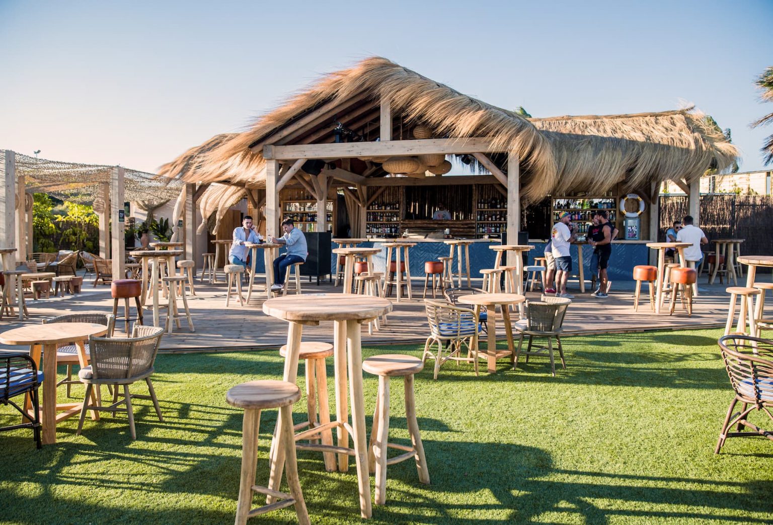 Chiringuito con cubierta de junco, mesas altas y césped, en Margarita Beach Club