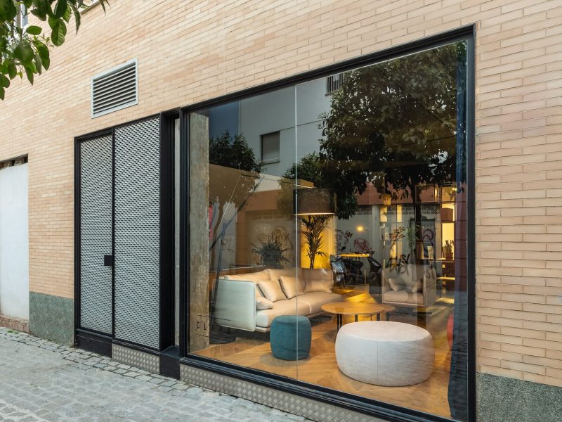 Fachada de ladrillo con gran escaparate y sofá blanco visible desde la calle