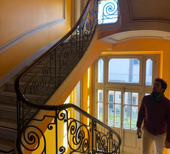 Escalera con barandilla de forja y paredes amarillas, con un hombre al pie