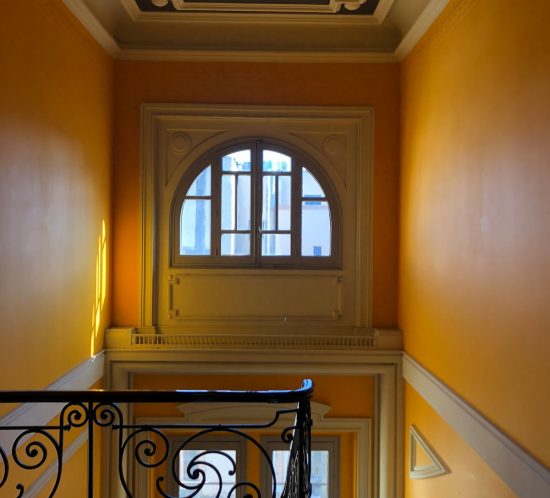 Caja de escalera con paredes amarillas, ventana en arco y barandilla de forja
