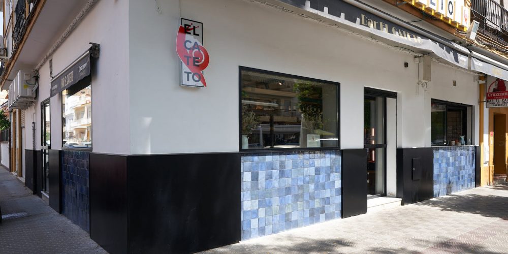Fachada en esquina de un bar con zócalo de azulejos azules y rótulo iluminado
