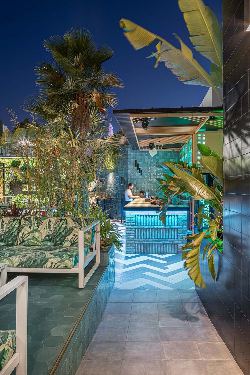 Terraza nocturna con palmeras, barra de azulejos y bancos tapizados con estampado de hojas