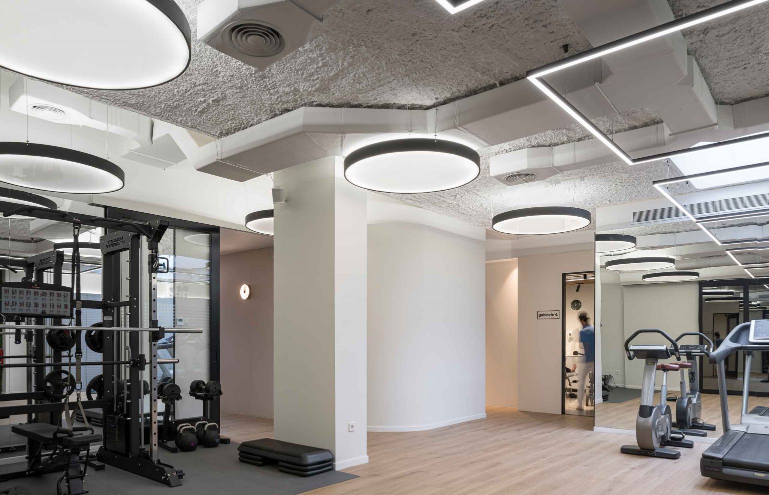 Sala de máquinas con techo de hormigón visto, luminarias circulares y bicicletas estáticas