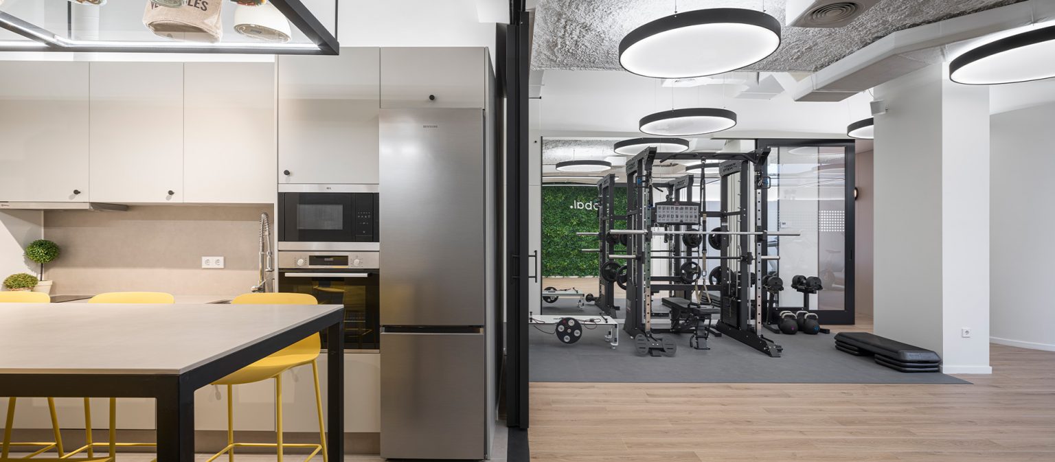 Cocina con taburetes amarillos abierta a sala de musculación con máquinas y pesas
