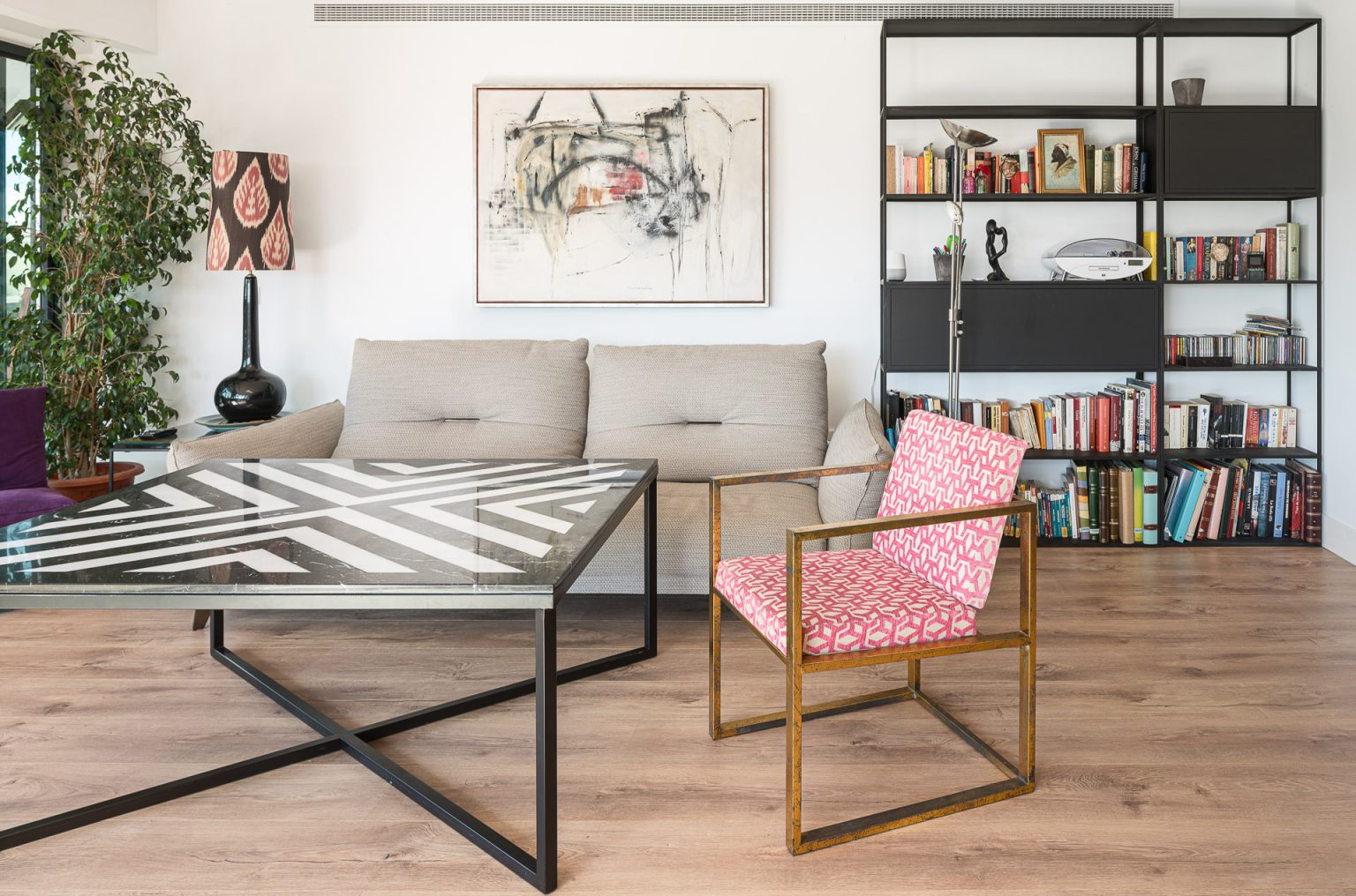 Salón con sofá claro, estantería metálica llena de libros y mesa baja de cristal