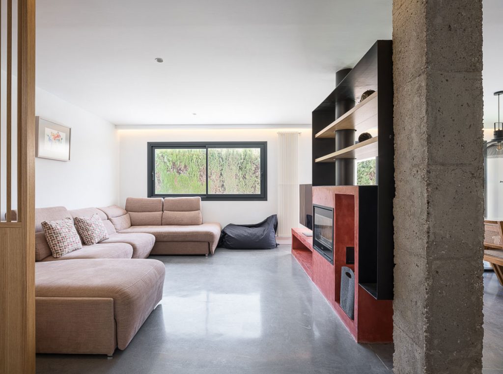 Infografía del salón con sofá en L, chimenea roja y pilar de hormigón visto