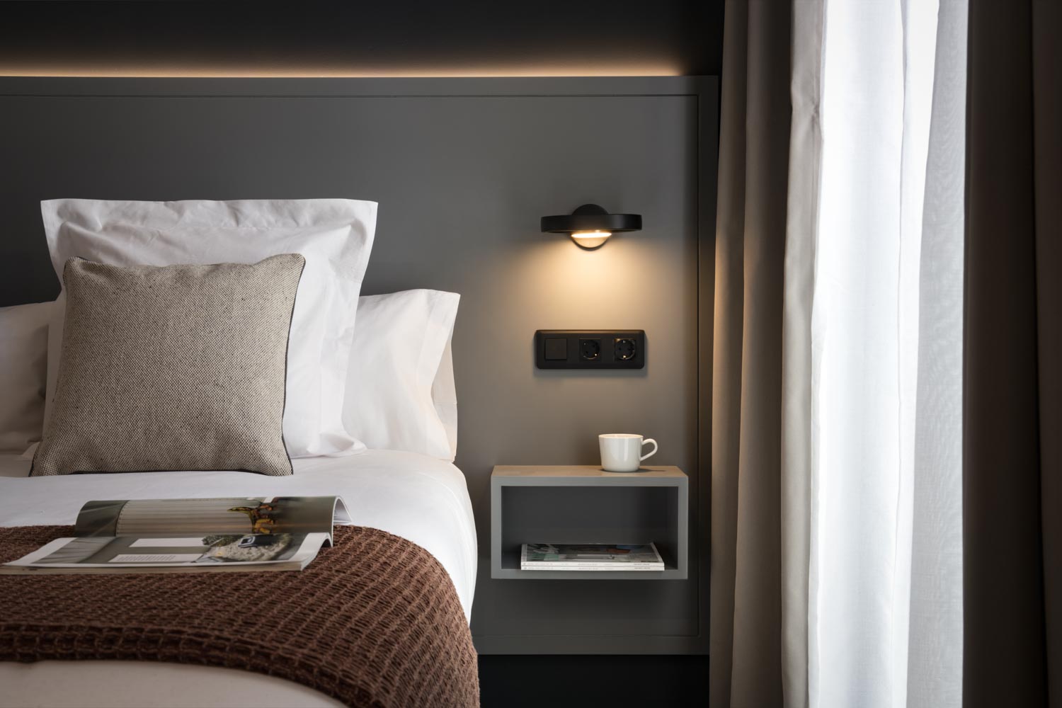 Cabecero gris oscuro con aplique negro, mesilla volada y cama con ropa blanca