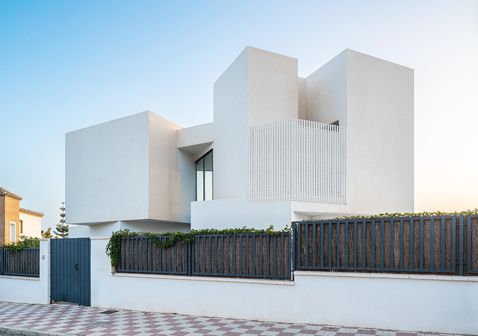 Fachada de volúmenes blancos con celosía vertical tras una valla oscura de listones