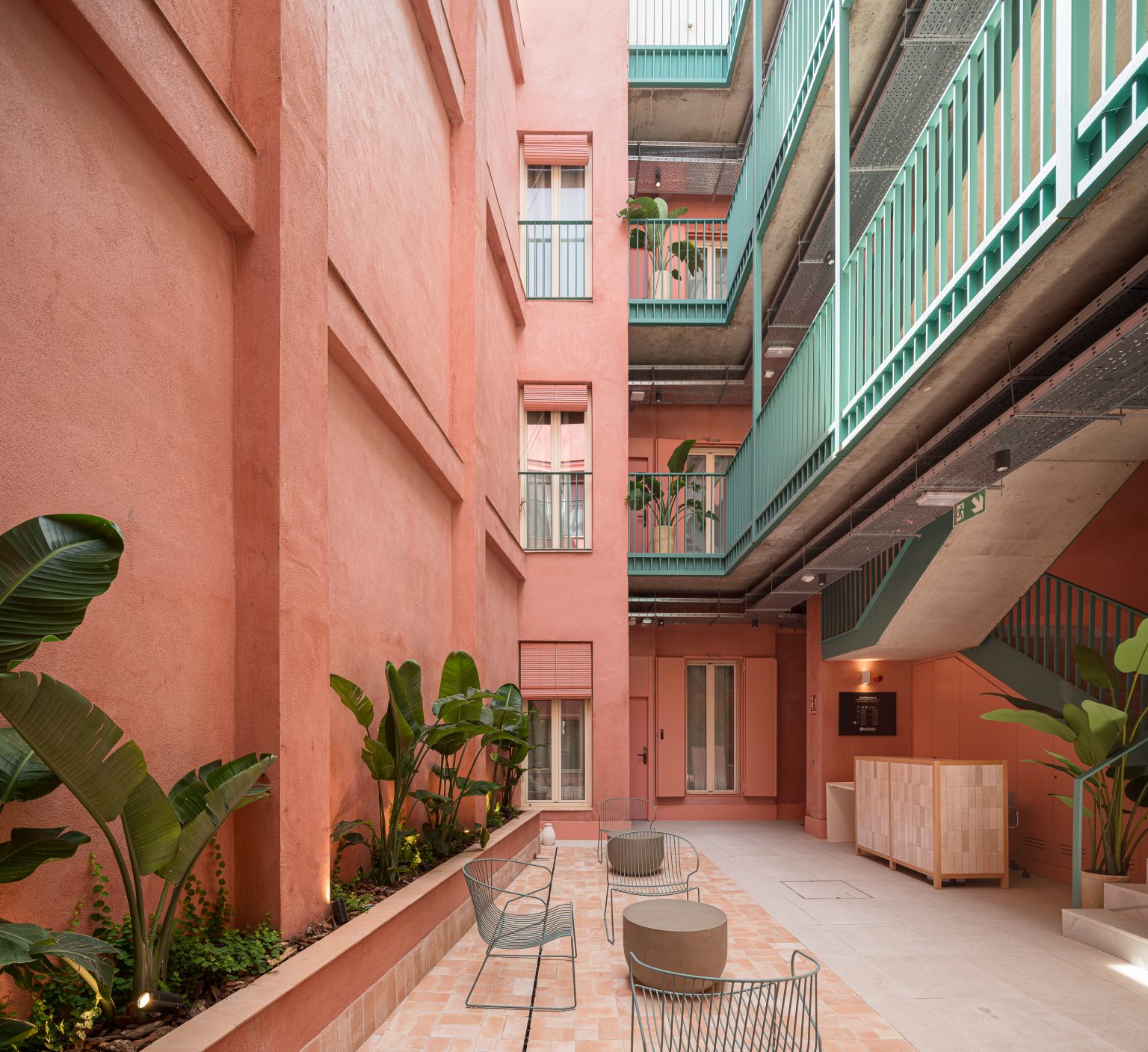 Patio con muros almagra, galerías verdes y sillas de rejilla en los apartamentos Clavellinas