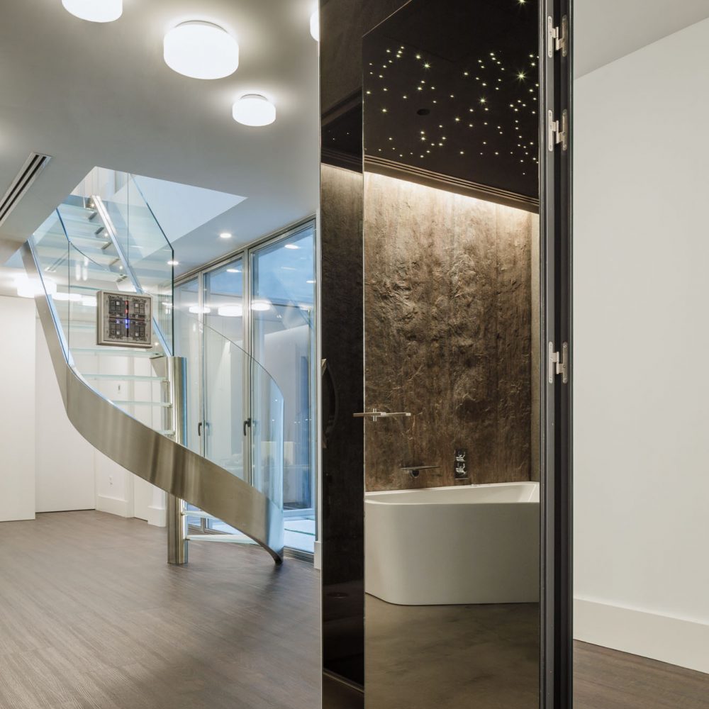 Escalera curva de acero y vidrio junto a baño con bañera exenta iluminada